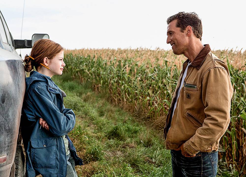 A imagem mostra cena do filme interestelar. É uma pai e uma filha, ela está encontrada na porta de um carro com as costas, olhando para seu pai, que por sua vez está com as mãos no bolso. Ao fundo, há uma enorme plantação.