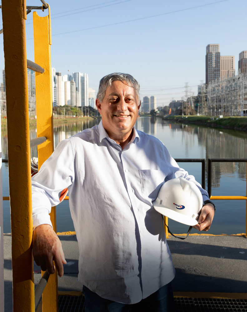 mergulhos-rio-pinheiros-itamar-emae-inversao-rio.jpg.jpg itamar sorrindo para a foto em plataforma sobre o rio pinheiros, apoiado em uma escada amarela e segurando o capacete branco no outro braço. ao fundo, o rio e prédios da cidade