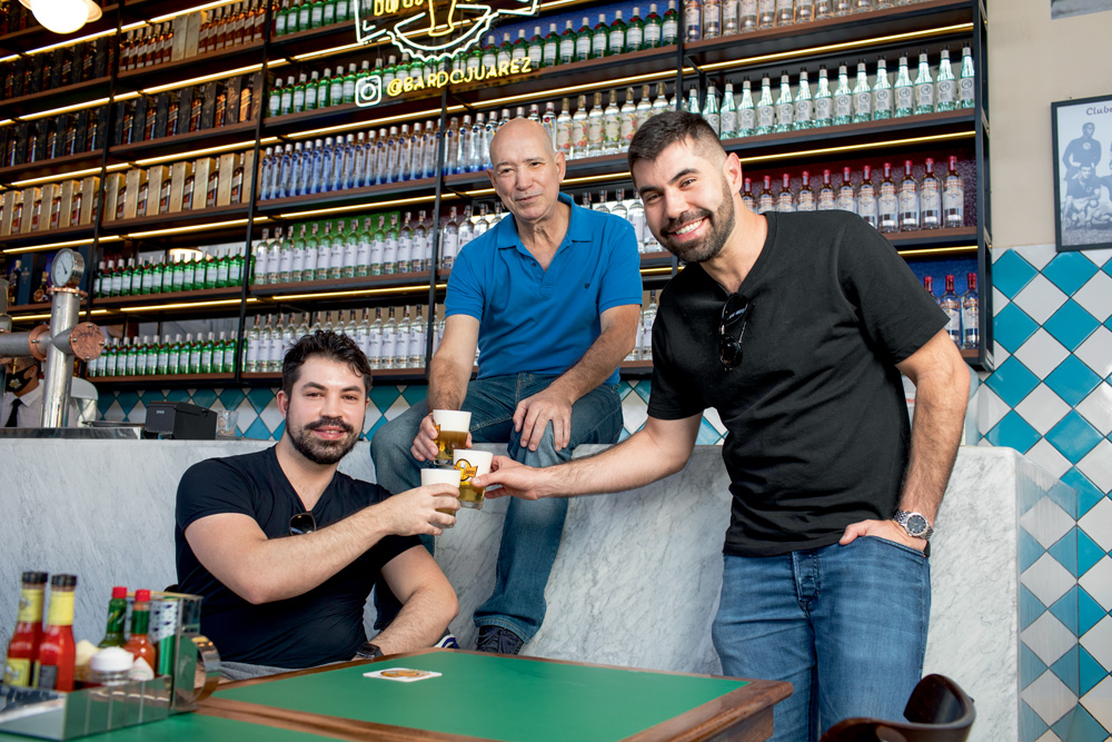 BARDOJUAREZlm14.jpg A imagem mostra Joarez, sentado no balcão do bar, e seus dos filhos ao lado, um sentado em uma cadeira e outro de pé, brindando copos cheios de chopp.