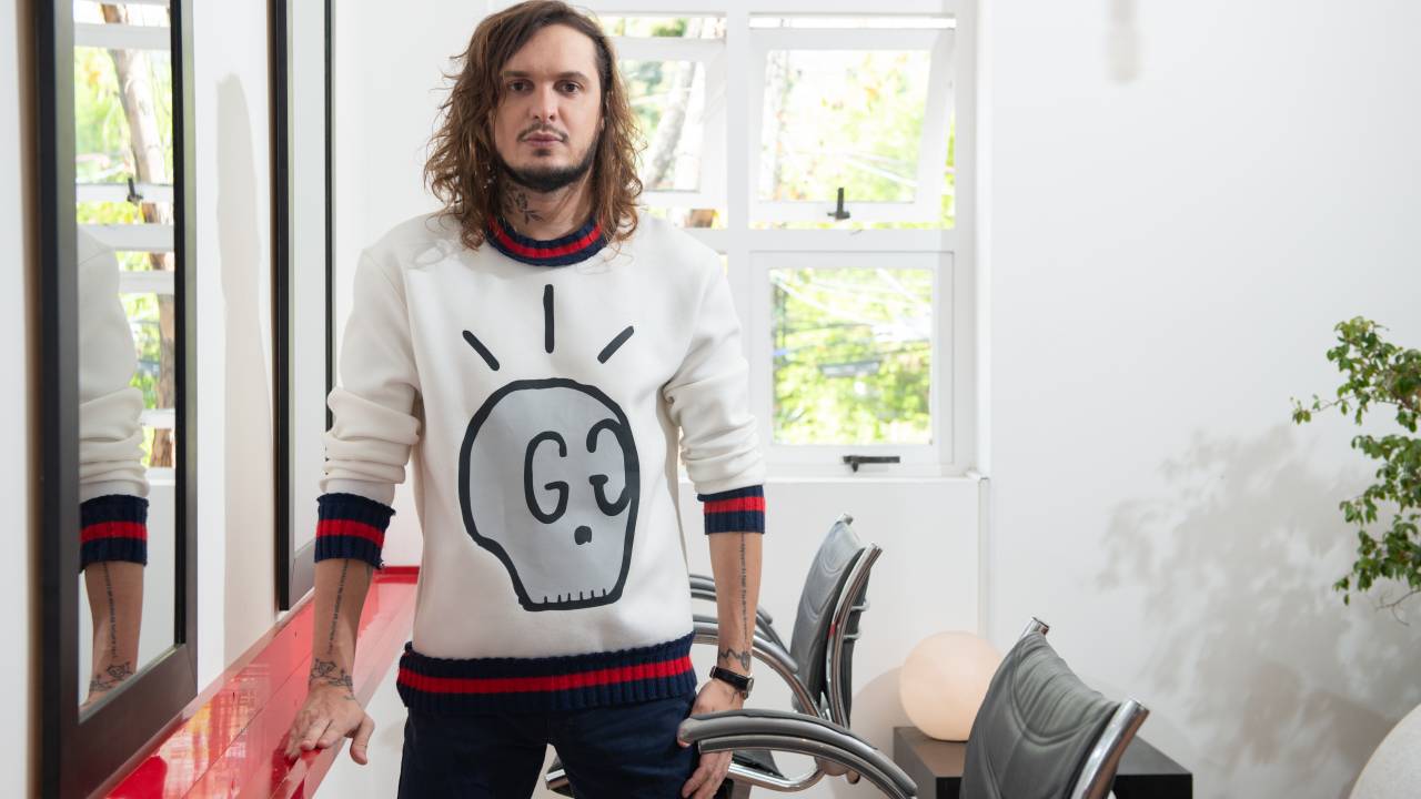 DJ Click posa de pé ao lado de mesa de salão de beleza. Veste suéter branco com figura em formato de caveira.