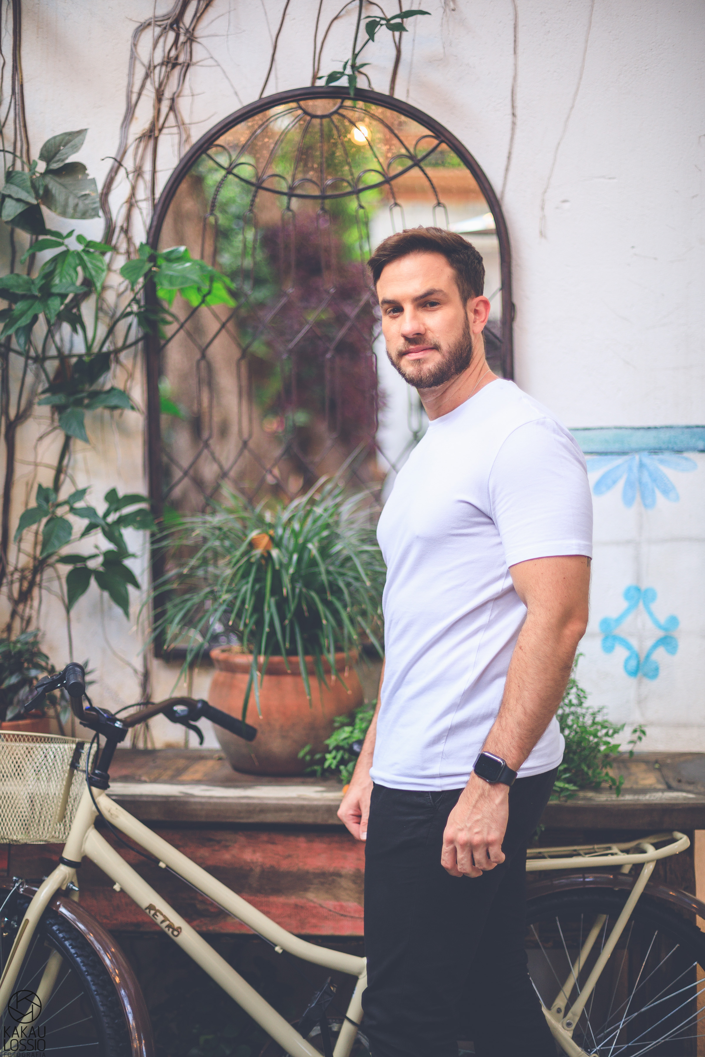 Daniel Adjuto posa de p&eacute;, de lado, olhando para a c&acirc;mera. Est&aacute; ao lado de plantas e uma bicicleta vintage. Veste cal&ccedil;a escura e camiseta branca.