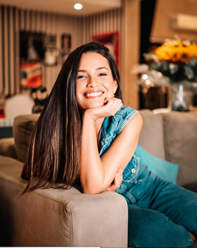Juliette Freire A imagem mostra Juliette sentada em um sofá, com a mão no queiro e sorrindo para a câmera.