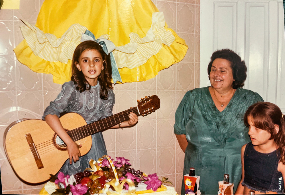 aniversário 7 anos com a avó Ivony e prima Daniela A imagem mostra Ludmilla com 7 anos, em seu aniversário, tocando violão enquanto é observada pela avó.