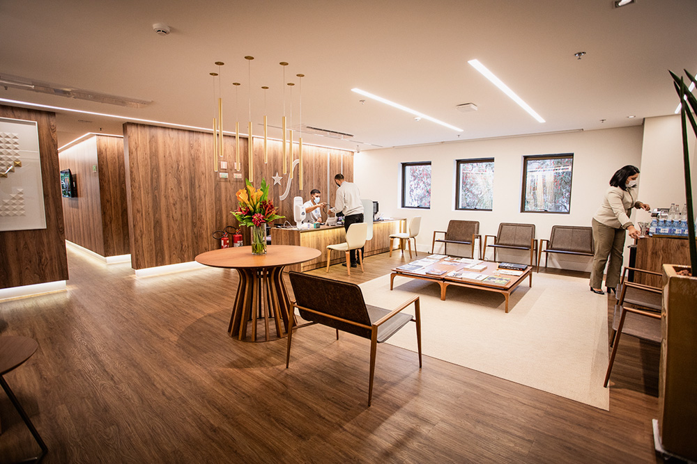 Ludhmila Hajjar sala de espera A imagem mostra um sala de espera larga. Há algumas cadeiras e a decoração é majoritariamente de madeira. Há duas pessoas na imagem, distantes, usando máscara.