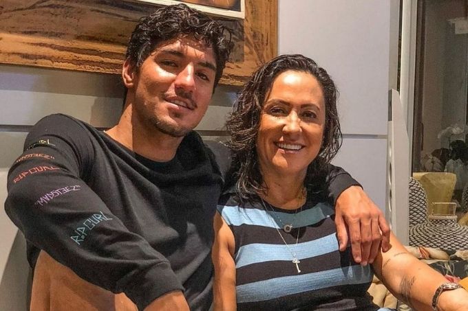 gabriel medina e simone medina sentados sorrindo para a foto. eles estão em casa, com suas costas encostadas em uma parede, e ele está com o braço em volta do pescoço dela