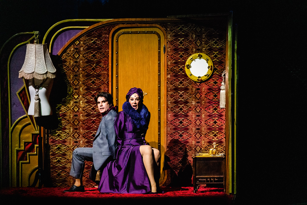 Em cena em um palco de teatro, Claudia Raia e Jarbas Homem de Mello estão sentados em um banco, de costas um para o outro, e com as mãos acorrentadas. Eles aparentam preocupação. Figurinos e cenário dos anos 1940.