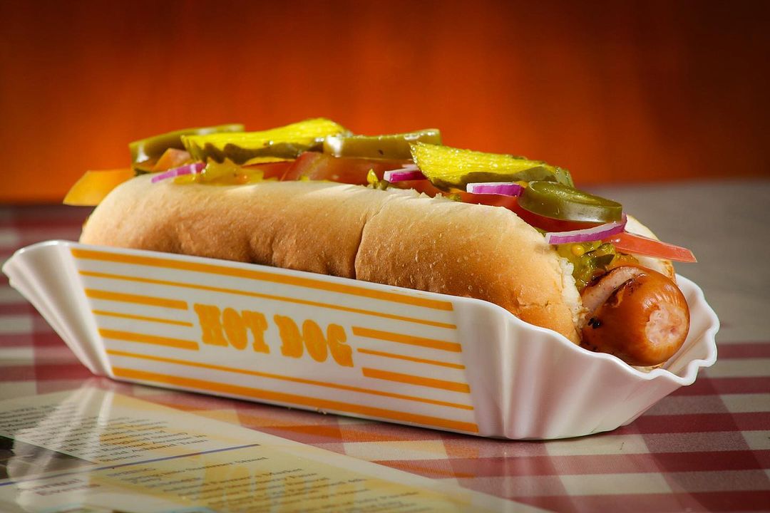 Sobre uma mesa coberta por uma toalha de flanela, o cachorro-quente está sendo servido em uma embalagem branca com um escrito (HOT DOG) e listras em amarelo. No cachorro-quente, em um pão longo, há à vista uma salsicha, tomate, cebola-roxa crua, e rodelas de pimenta jalapeño.