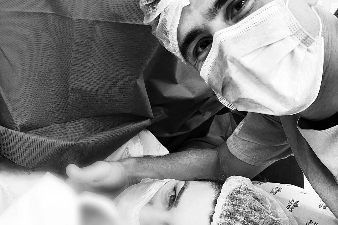 A imagem mostra o jogador Willian Bigode e a esposa Loisy em sala de cirurgia; ela está deitada com o bebê nos braços