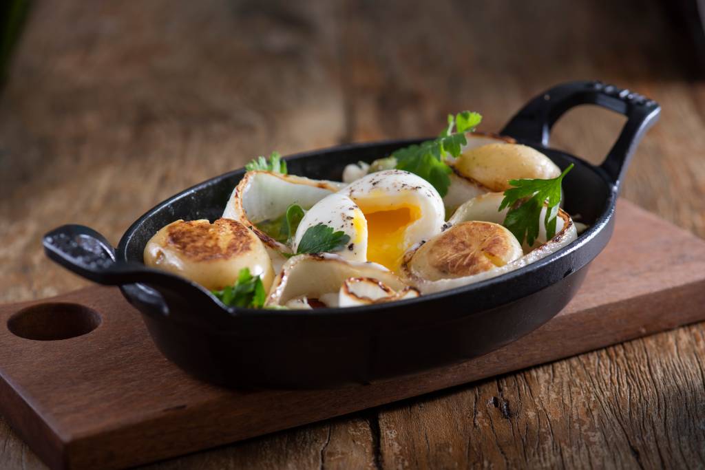 COMER & BEBER 2021 – Chez Claude Assadeira de ferro preto com bacalhau, ovo mole e batatas ao murro sobre bancada de madeira.