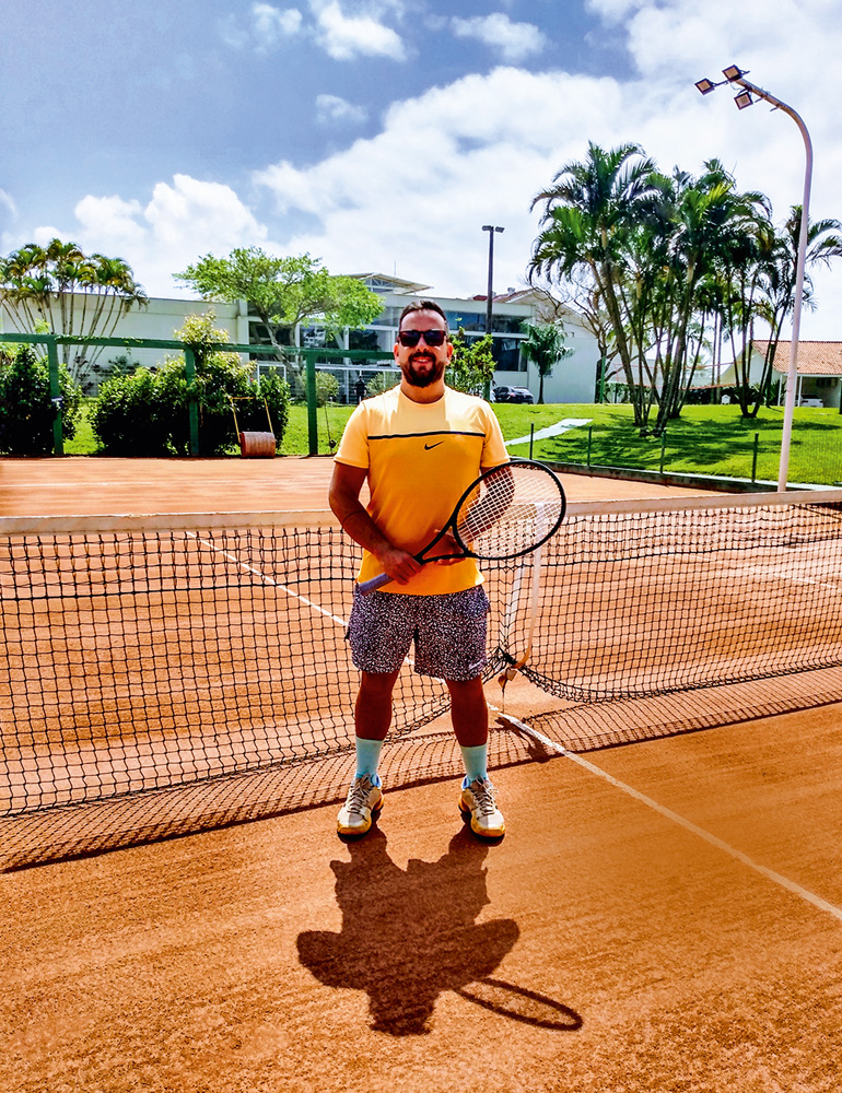 Guilherme GRAAC_3 A imagem mostra Guilherme, já adulto, em uma quadra de tênis, segurando uma raquete de pé em frente a rede. Ele está de óculos escuro.