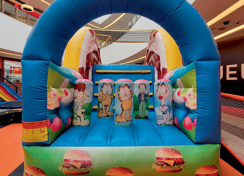 Em um espaço dentro de um shopping, há um brinquedo inflável decorado com imagens do personagem Garfield. Há um espaço para pular e escorregar