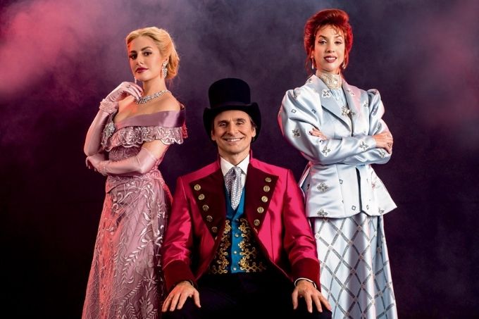 murilo rosa, sentado, sorrindo para a foto vestido de showman com terno vermelho e chapéu. ele está acompanhado de duas mulheres, Giulia Nadruz e Diva Menner, que estão de pé