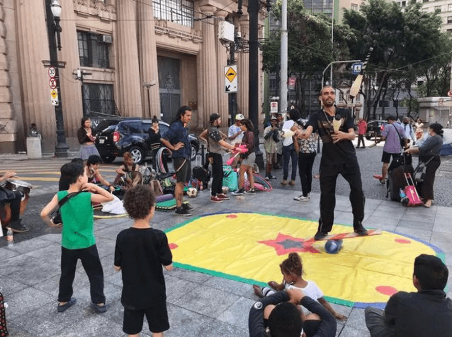 sebastião Em uma praça, há crianças em volta de um circulo com um tapete colorido de brincar. Há um homem fazendo malabarismos no centro da roda