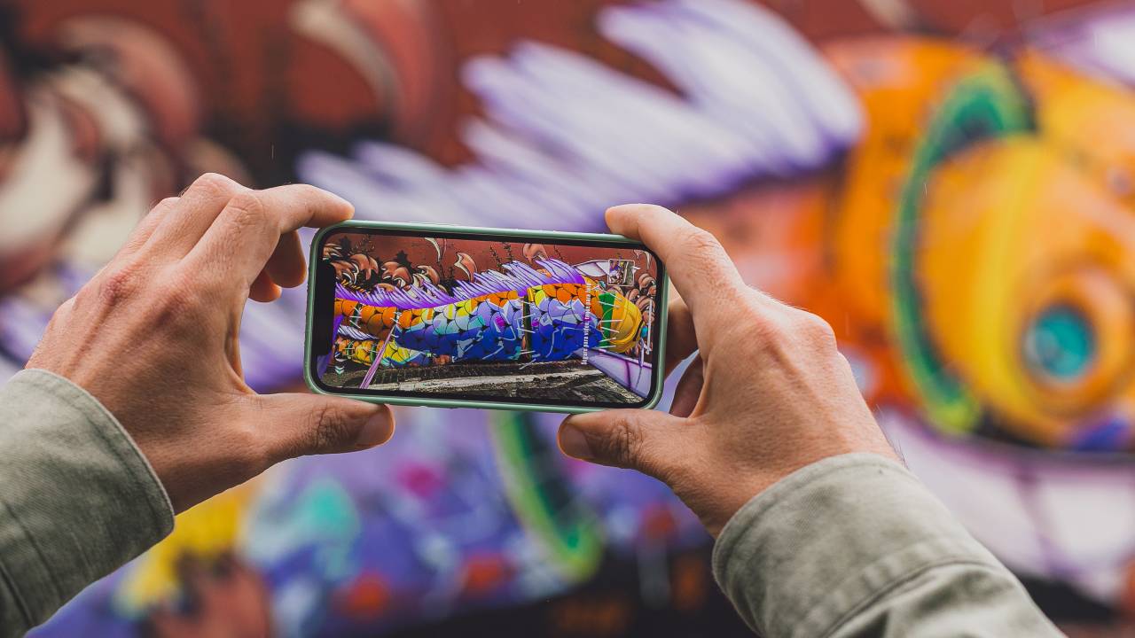 Tela de celular apontada para um grafite, que surge em realidade aumentada. É uma obra no Beco do Batman