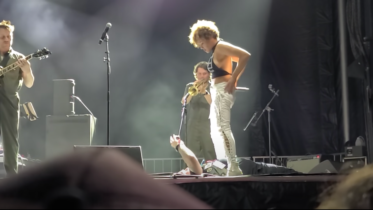 Foto do palco de show, mostra um guitarrista do lado esquerdo e, no centro da imagem, uma mulher em pé em cima de um homem, prestes a abaixar sua calça branca