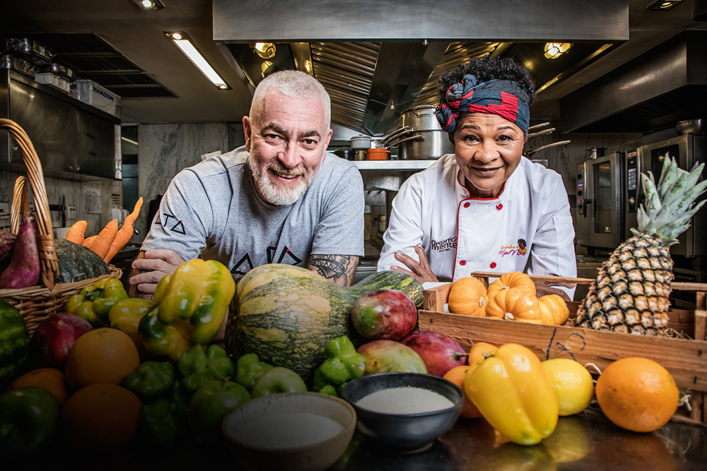 Em uma cozinha, Alex Atala posa ao lado de Tia Nice junto de frutas e legumes espelhados na frente da dupla