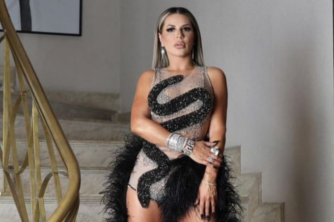 Mulher com vestido transparente com detalhes em preto em pé sobre uma escada de mármore e corrimão dourado.