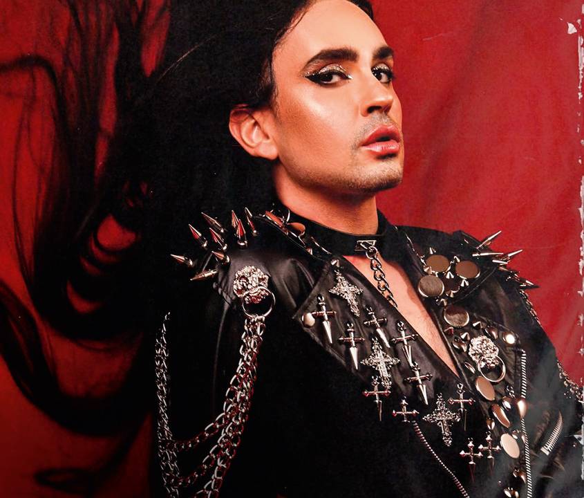 A imagem mostra Johnny Hooker apoiado em uma parede vermelha e encarando a câmera.