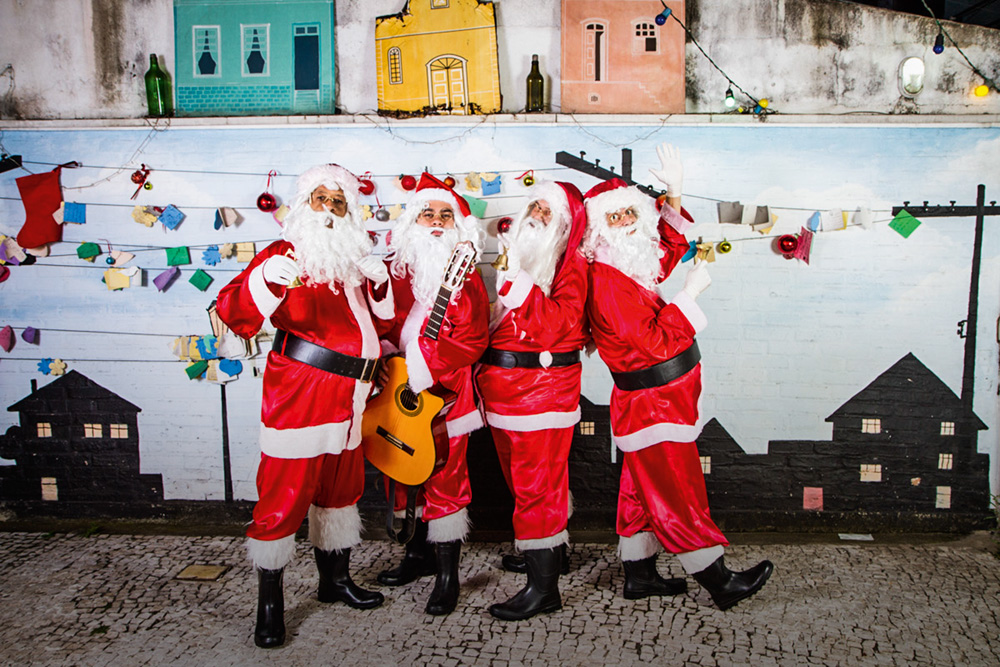 Em um cenário de Natal, quatro pessoas vestidas de Papai Noel posam juntas. Um segura um violão