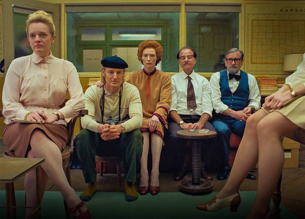 Imagem mostra 6 pessoas sentadas, 4 em um sofá no centro. Todos vestem roupas formais retrô