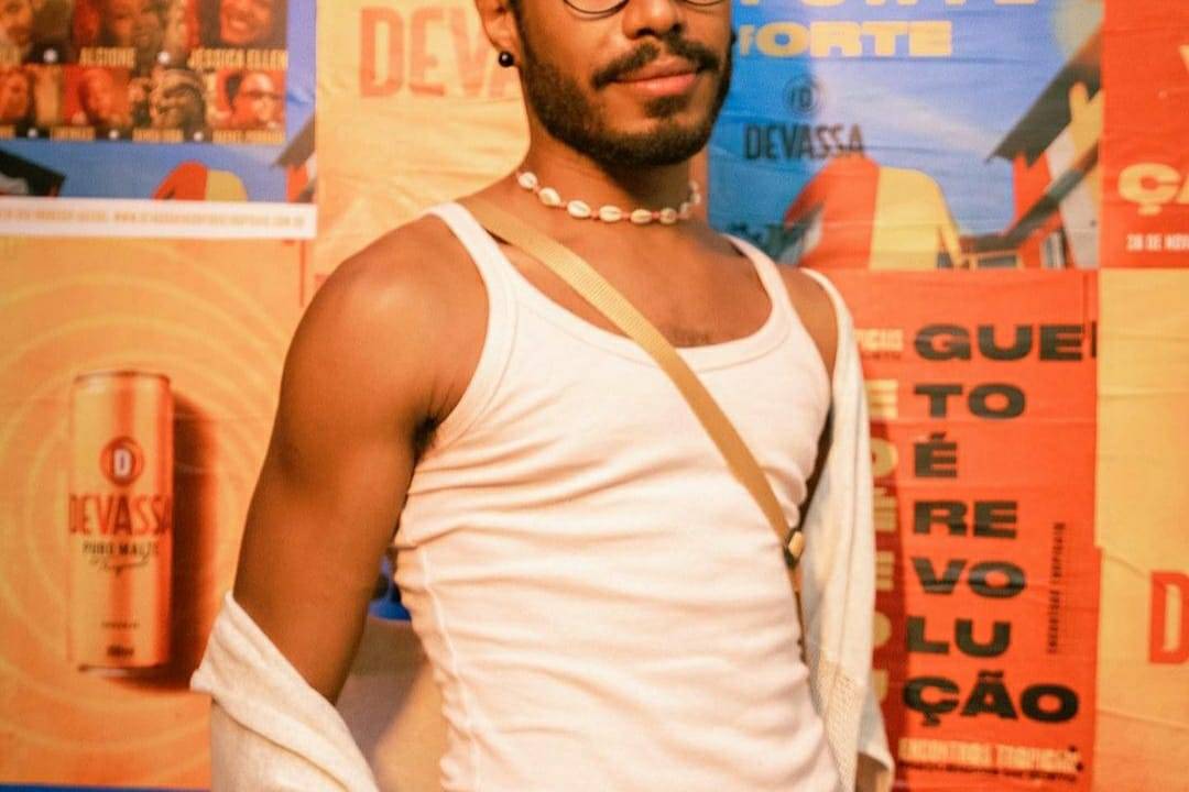 Foto de Ícaro Silva. Ele é um homem negro, de cabelos crespos, olhos e barba escuros. Usa óculos e veste um conjunto branco de regata, calça e camisa. Ele também usa um colar e leva uma bolsa transpassada pelo corpo.