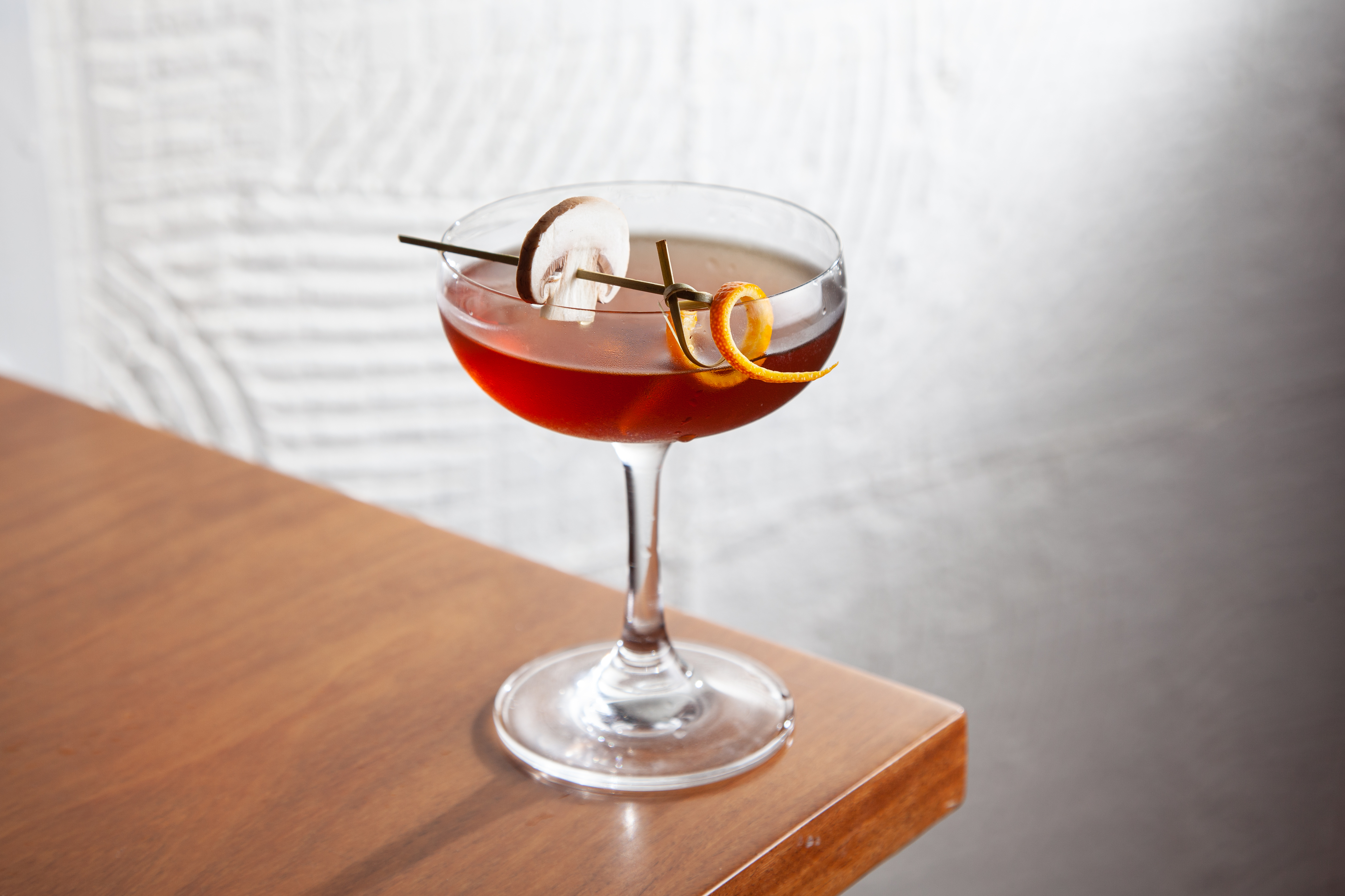 Ta&ccedil;a coupe com um drinque de cor alaranjada e decorada com um peda&ccedil;o de casca de laranja e uma lasca de cogumelo