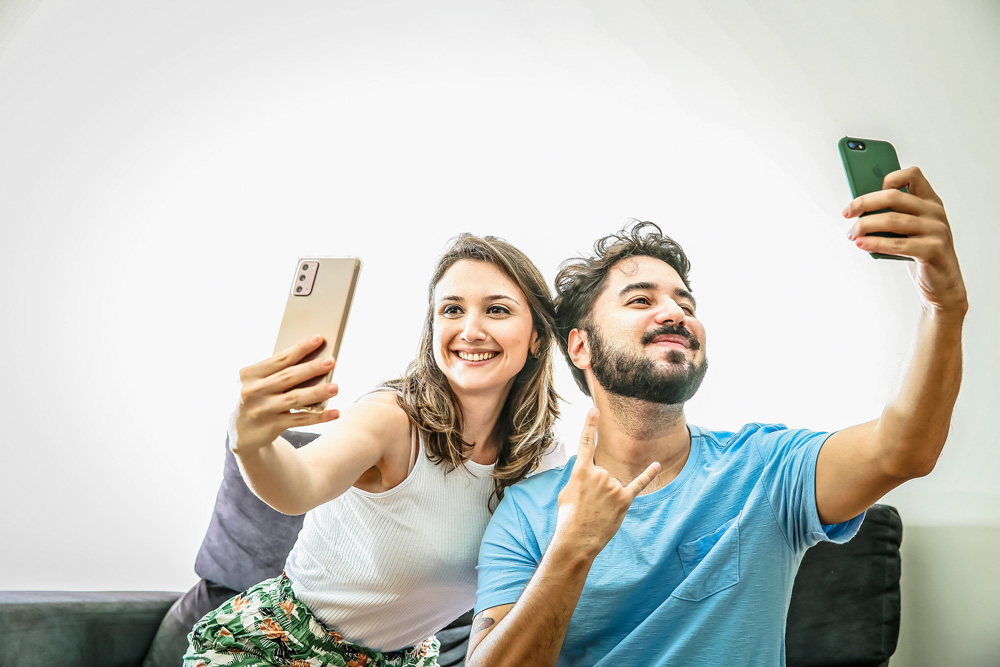 CASALAPP_MARIANA_6.JPG Um casal está sentado no sofá. Ambos possuem um celular em suas mãos e posam para uma foto em cada um dos smartphones