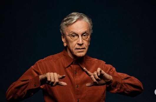 Imagem mostra CAetano Veloso vestindo camisa marrom e com os dedos indicadores e polegares, das duas mãos, levantados