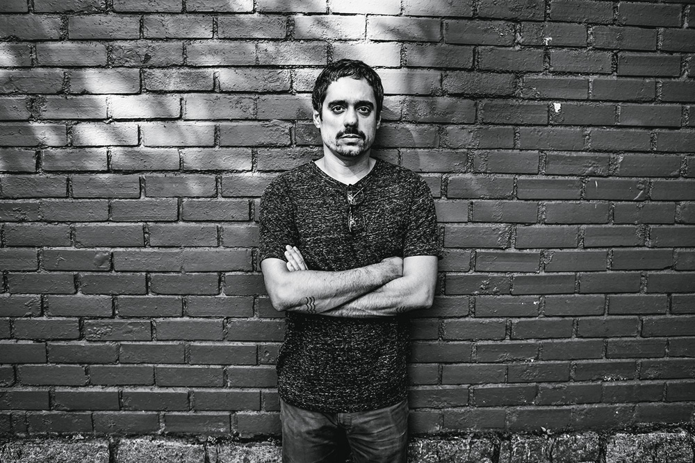 Produtor musical Chico Dub posa encostado em parede de tijolos com os braços cruzados. Tem barba e cabelo curto. Foto em preto e branco.