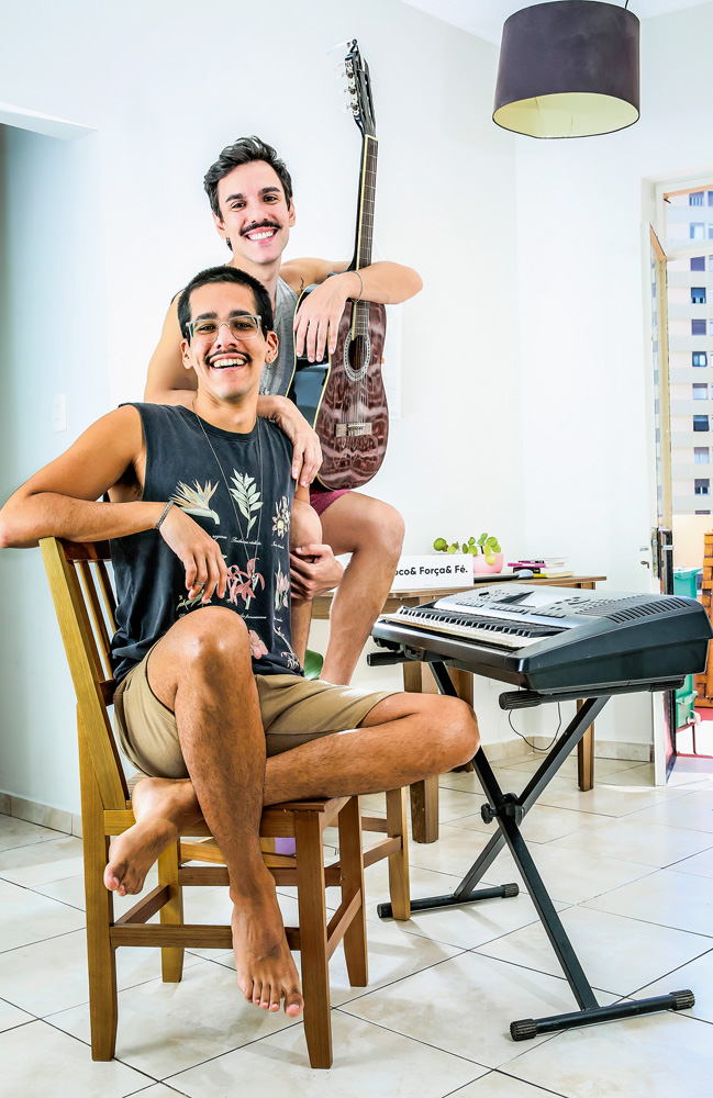 São Paulo 10 dezembro 2021Rafael e Victor.Foto Alexandre Battibugli VSP Um casal posa sorrindo para foto. Um deles está sentado em uma cadeira e em frente a um piano e o outro está de pé, atrás da cadeira, segurando um violão