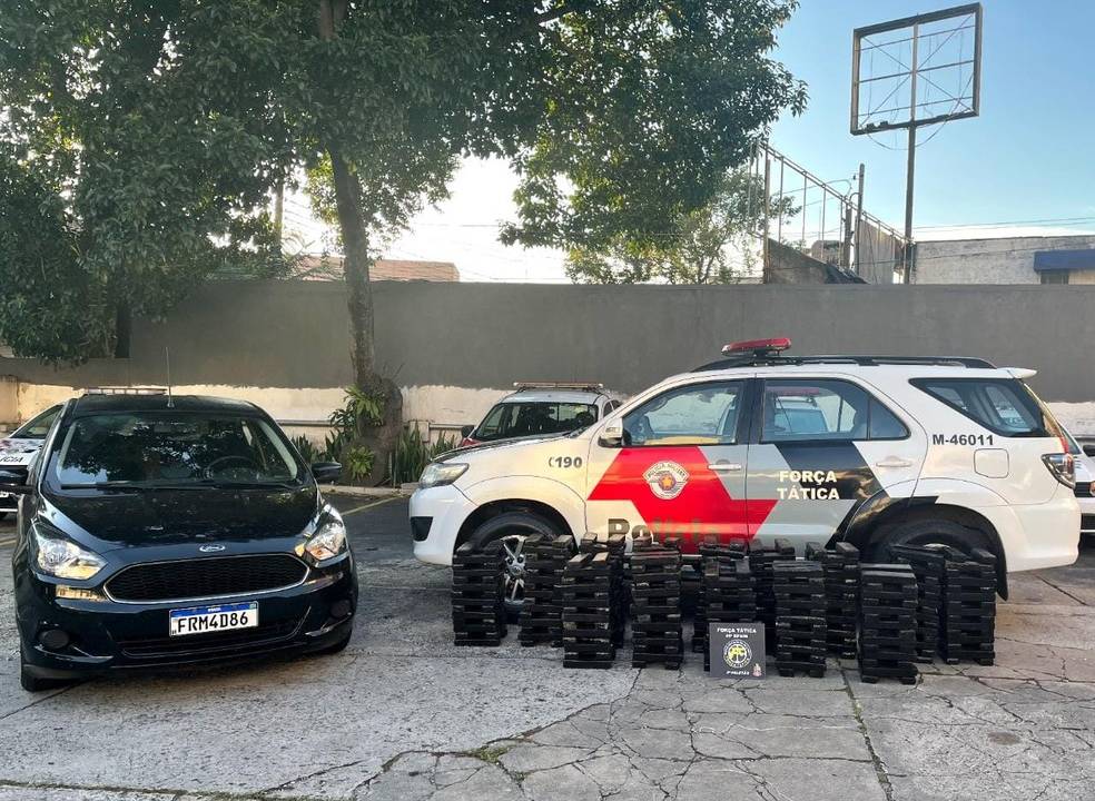 Foto mostra carro preto e viatura estacionados com carregamento de maconha disposto no chão.