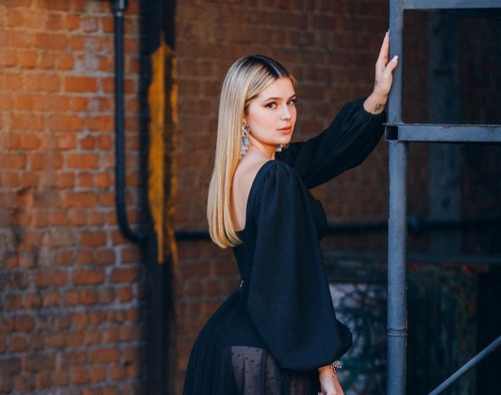 Imagem mostra mulher loira de vestido preto com a mão apoiada em uma parede, olhando para a câmera.