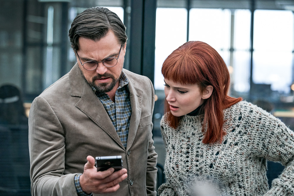 Imagem mostra um homem de paletó segurando um celular ao lado de uma mulher ruiva que olha para a tela com uma cara de susto.