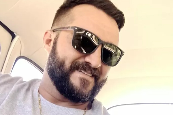 Imagem mostra homem de óculos escuros, barba e cabelo curto, usando camiseta e corrente de ouro, sorrindo