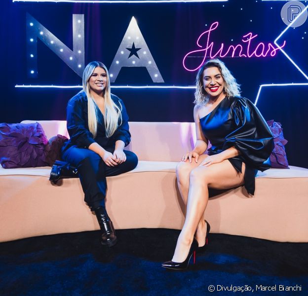 Imagem mostra duas mulheres de roupas pretas sentadas em sofá branco com almofadas roxas