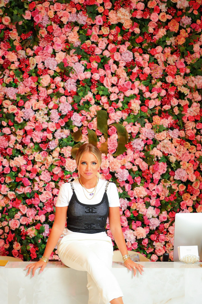 Ateliê Beauty.jpg Imagem mostra mulher loira de roupa branca e preta sentada em frente a paredão de rosas da cor rosa e vermelha.