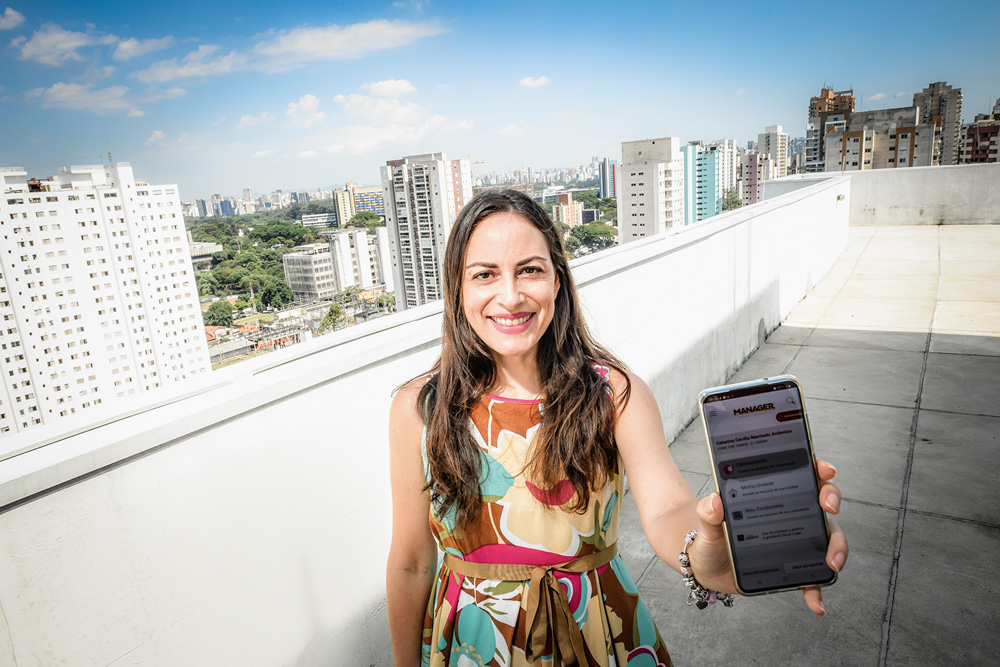São Paulo 22 dezembro 2021Foto Alexandre Battibugli VSP Catarina Anderáos aparece mostrando seu telefone celular para a câmera, em uma sacada de um condomínio. Ela é uma mulher branca, de cabelos e olhos castanhos e usa um vestido colorido e estampado.