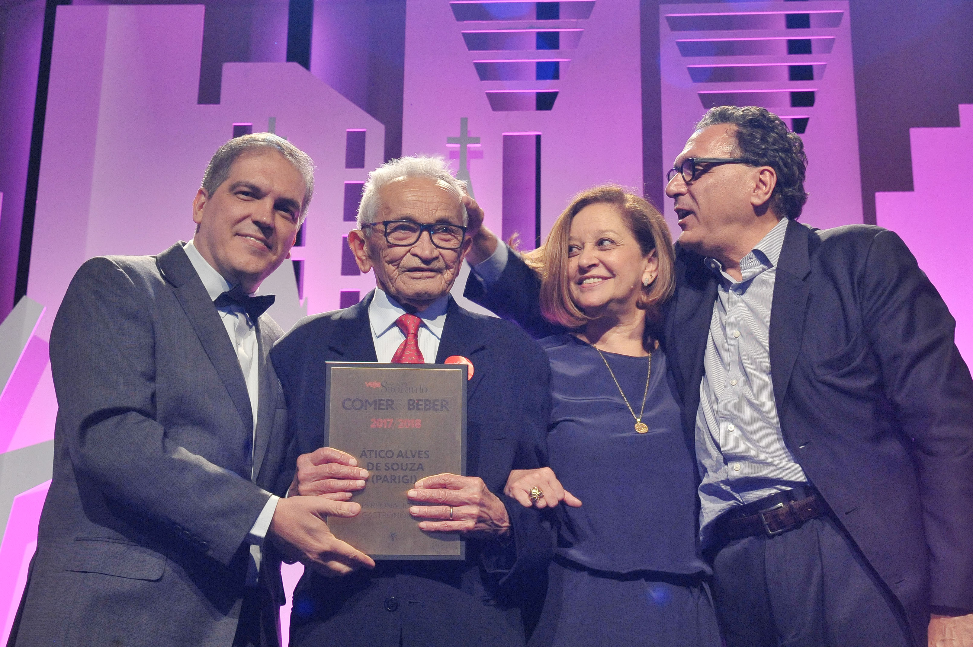 Homem idoso segura placa. &Agrave; direita dele, homem de grava borboleta. &Agrave; esquerda dele, mulher e homem.