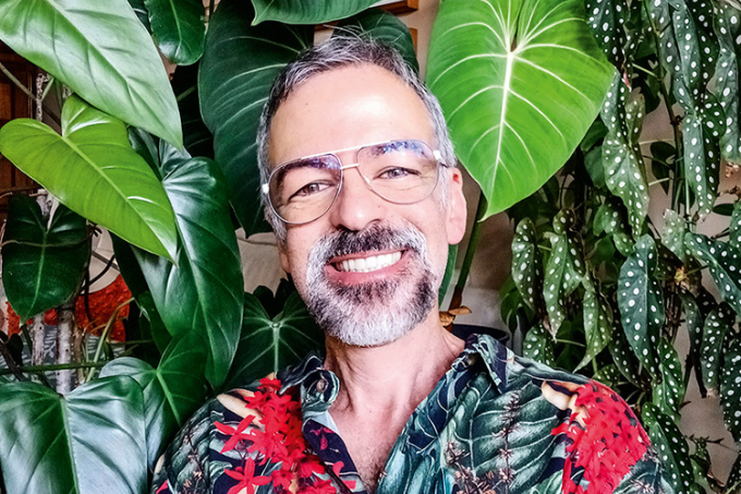 Imagem mostra homem de camisa florida em frente a parede com diversas plantas penduradas.