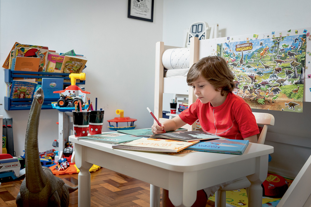 Gabriel_6_foto_Wanezza_Soares-0160-Edit.jpg Gabriel aparece sentado em uma mesa branca. Ele escreve em um caderno. O ambiente ao redor é decorado com brinquedos.