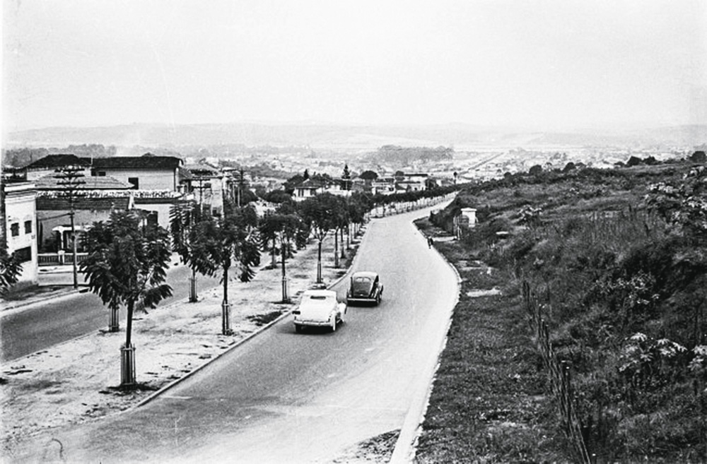 Reboucas-no-sentido-centro-bairro-fotografia-de-benedito-junqueira-dua-e1540493529482.jpg Imagem em preto e branco mostra rua, ao centro, com dois carros trafegando. À direita da rua, mato, à esquerda, casas.
