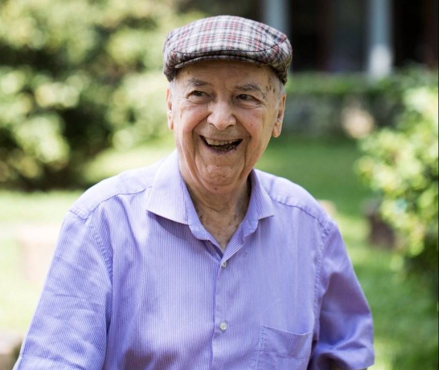 Imagem do padre sorrindo. Ele veste uma camisa azul claro e usa boina com estampa xadrez