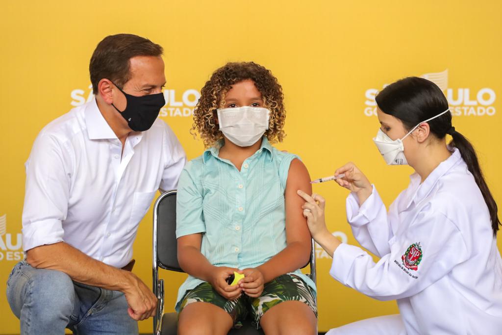 Imagem mostra criança sendo vacinada por enfermeira ao lado de João Doria