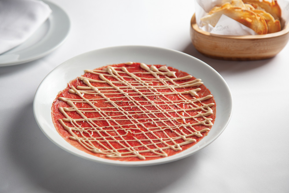 Carpaccio servido em prato raso de louça branca junto de porção de pães servida em recipiente de madeira