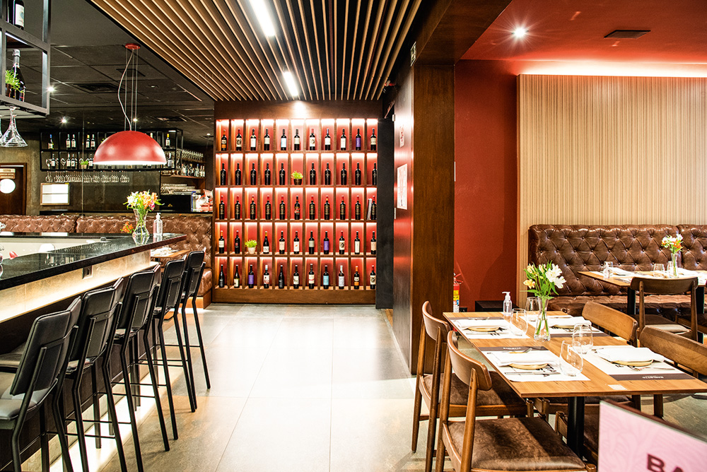 Ambiente do restaurante com mesas decoradas por pequenos vasos com flores sobre elas, sofás de couro marrom contra as paredes, balcão com banquetas pretas e adega ao fundo