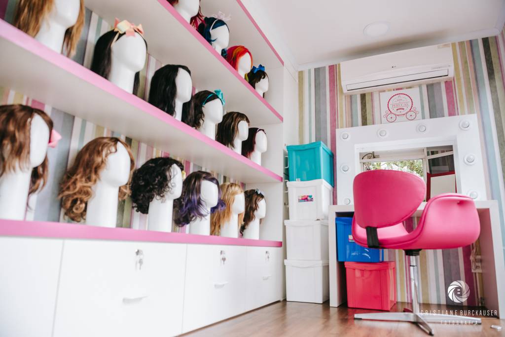 Cabelegria 07 Foto mostra o interior de uma caminhão. Quatro prateleiras cor-de-rosa abrigam suportes para perucas. Ao lado, há uma penteadeira, uma cadeira giratória também rosa e um espelho para provar as perucas.