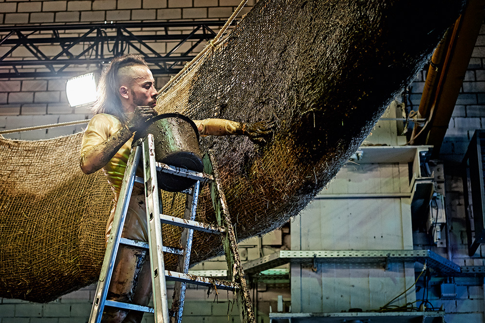 Daniel Lie, perfil – Credits Martin Meiser. Imagem mostra pessoa em escada prata com balde de tinta, passando em estrutura cilíndrica de juta com feno.