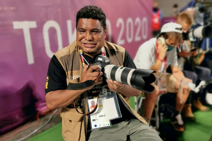 Imagem mostra homem segurando câmera, sorrindo. Ao fundo, uma faixa escrito "Tokyo 2020"