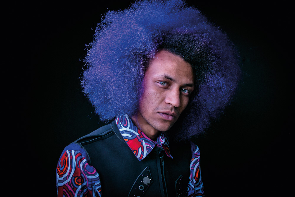 Edgar_foto Camila Rhodes.jpg Imagem mostra homem com cabelo iluminado por luz roxa, vestindo camisa e colete preto.