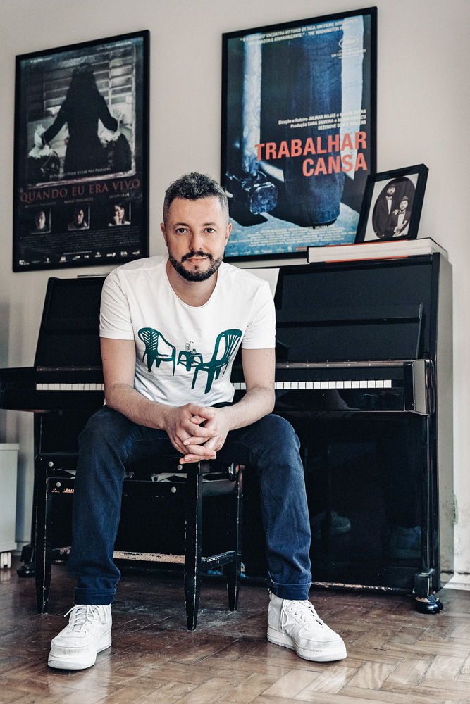 Marco Dutra Imagem mostra homem de camseta branca e calça jeans sentado em banco à frente de um piano. Na parede, pôsteres de filmes.